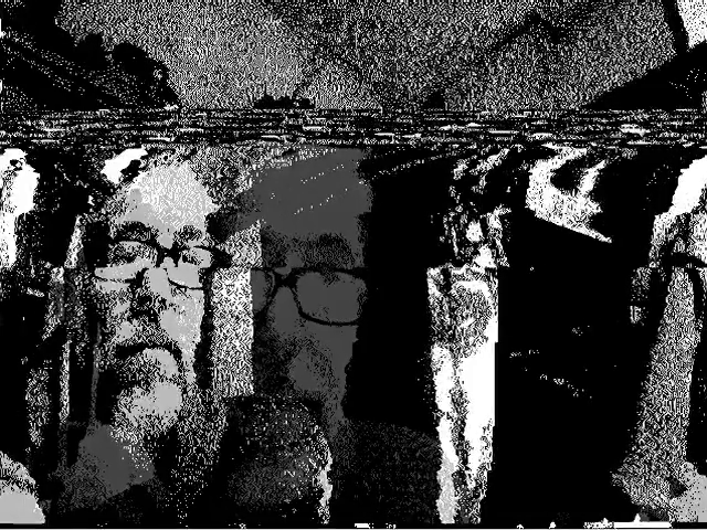 A grainy black and white image, similar to TV static or radio noise when put on an empty channell. In the static there is man's face fainly visible. He wears square reading glasses with a thick frame. His face is visible in several instances over the frame, but each instance is in some way distorted, as if melted wax.