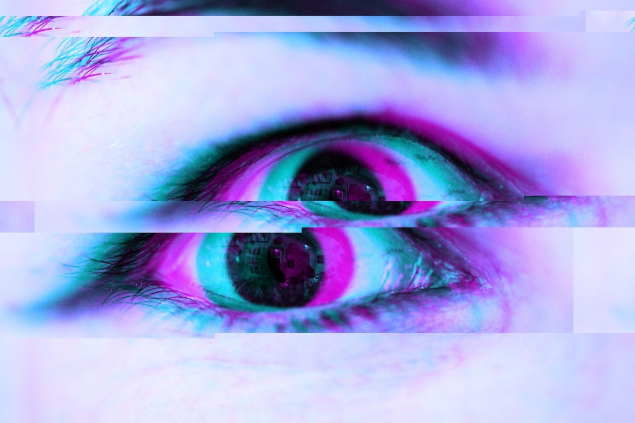 An image of an eye. The image is digitally manipulated so the eye seems fractured horisontally into two eyes, both draped in hues of purple and mint green. There is a faint but recognizable reflection of a brick building with four windows in the pupil's reflection.